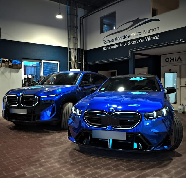 Blauer BMW XM und BMW M5 in der Halle, dahinter das Firmenlogo an der Wand
